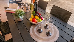 Une table à manger en plein air, agrémentée d’un livre, d’une plante en pot, d’une bouteille de vin, d’un saladier de fruits et de verres à vin.