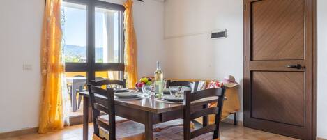 A dining area with a table set for two, chairs with cushioned seats, a window with curtains, and a door with a wooden frame.