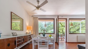 A dining area with a table and chairs, a ceiling fan, a window with curtains, and a view of trees outside.