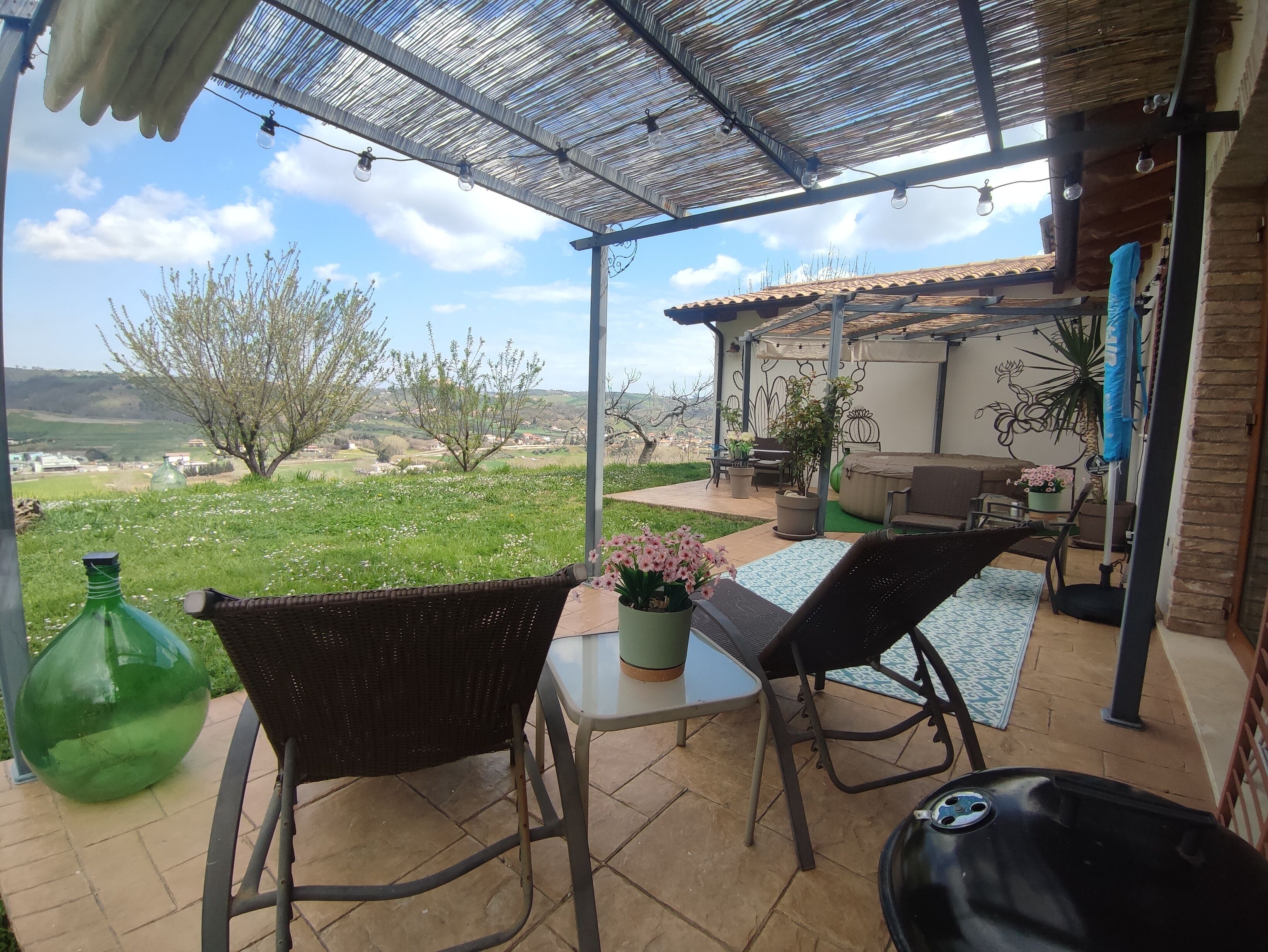 En overdækket terrasse med udendørs møbler, en grøn flaske og udsigt til landskabet.