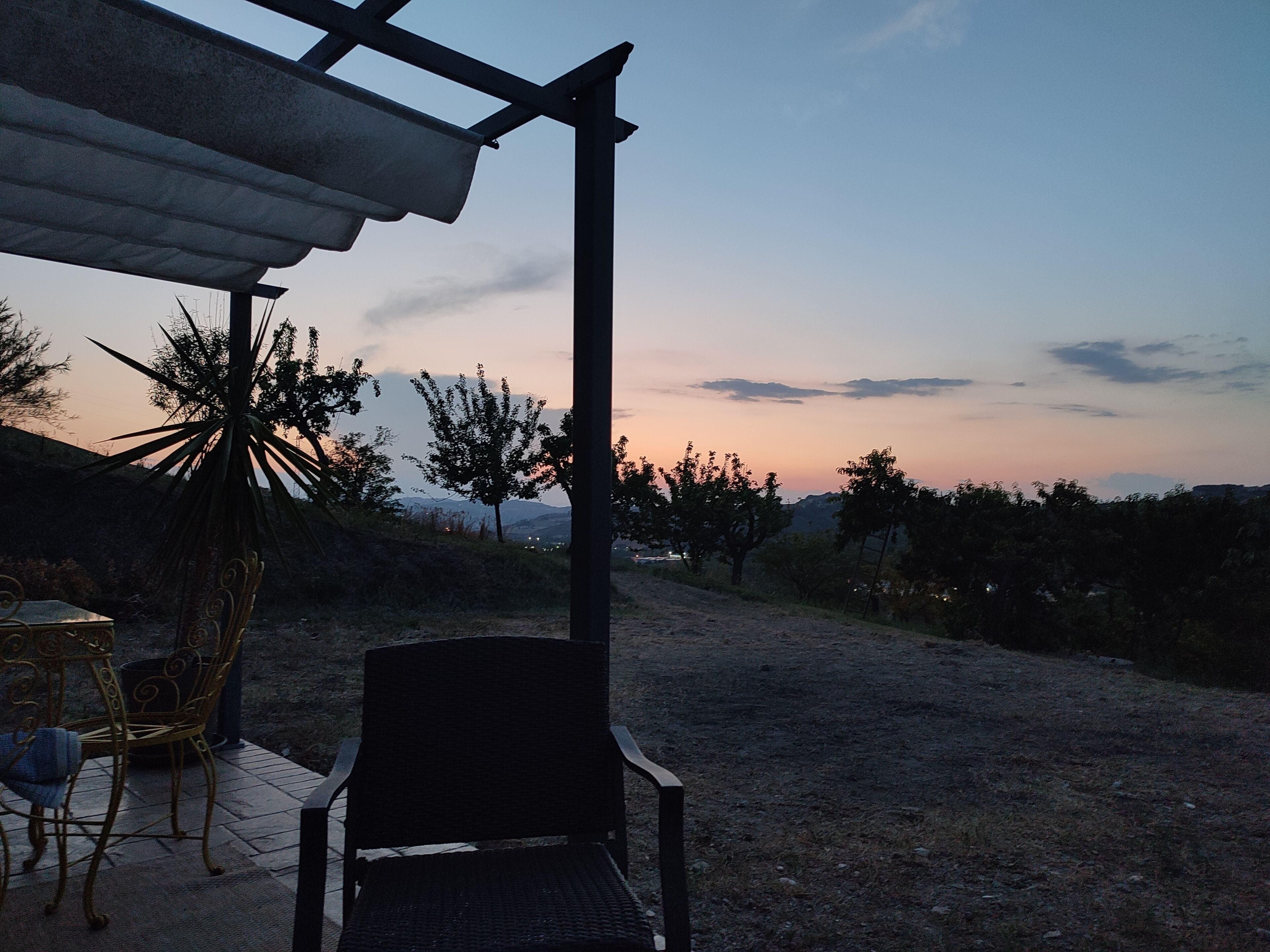 Un espace extérieur aménagé avec une chaise et une table, installé sous une pergola, d’où l’on peut admirer un paysage composé d’arbres et de montagnes lointaines.