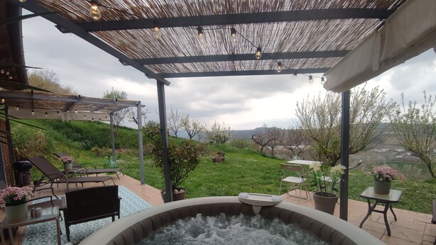 Een hottub met uitzicht op een gazon en bomen, een pergola met lichtslingers en tuinmeubilair.
