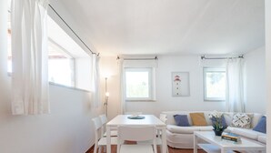 A bright living room with a white sofa, a dining table with chairs, and a coffee table. There are windows with curtains, a floor lamp, and a framed picture on the wall.