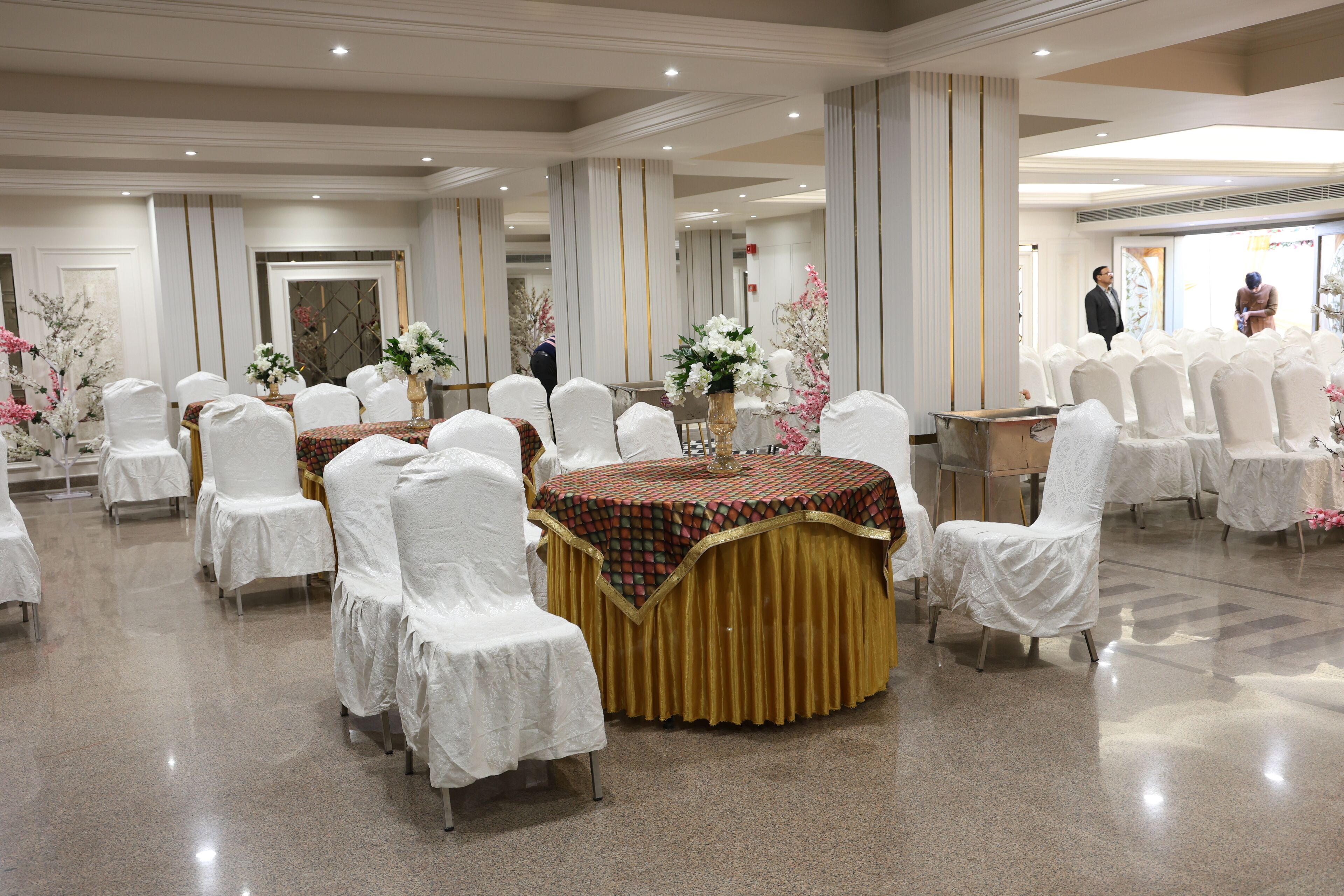 Uma sala de banquetes com mesas redondas cobertas por toalhas, cadeiras brancas e decorações florais.