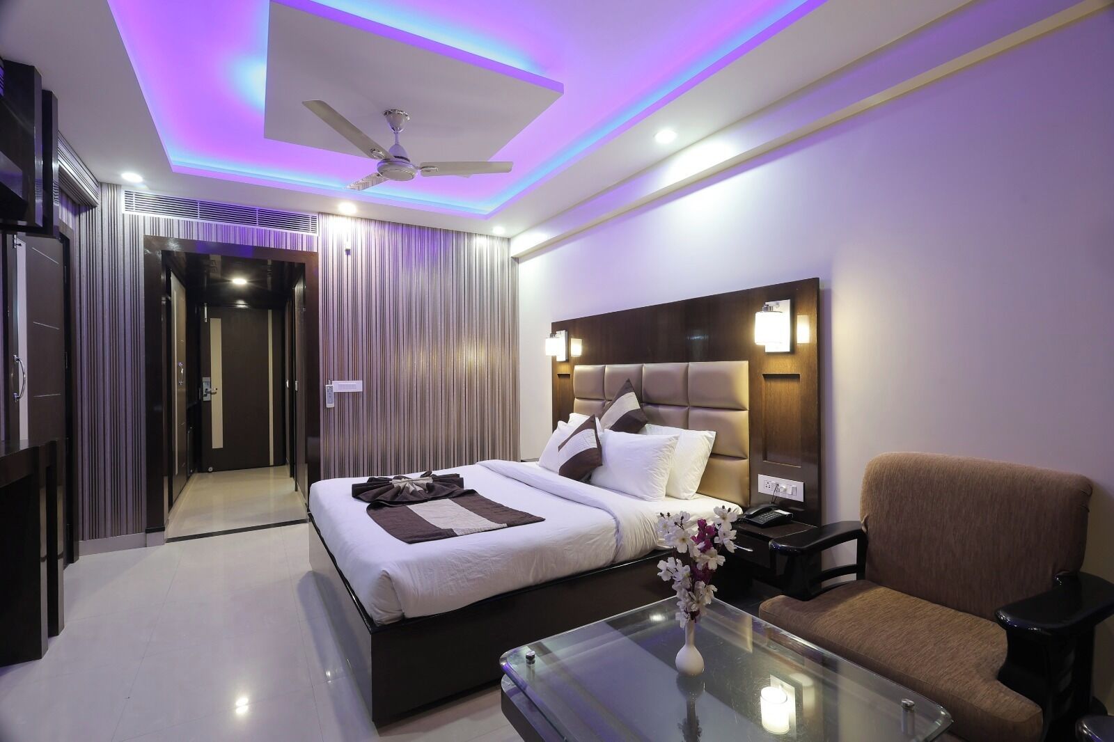 A modern hotel room with a bed, a chair, a glass table, and a ceiling fan.