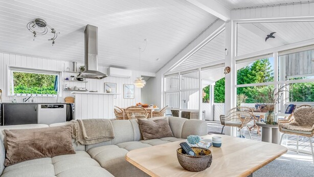 A spacious living area with a large sofa, a wooden coffee table, and wicker chairs. There is a kitchen area with a refrigerator and a window overlooking greenery.