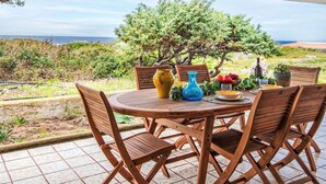 Un tavolo rotondo in legno con quattro sedie, su un patio piastrellato con vista su un paesaggio costiero, alberi e il mare.