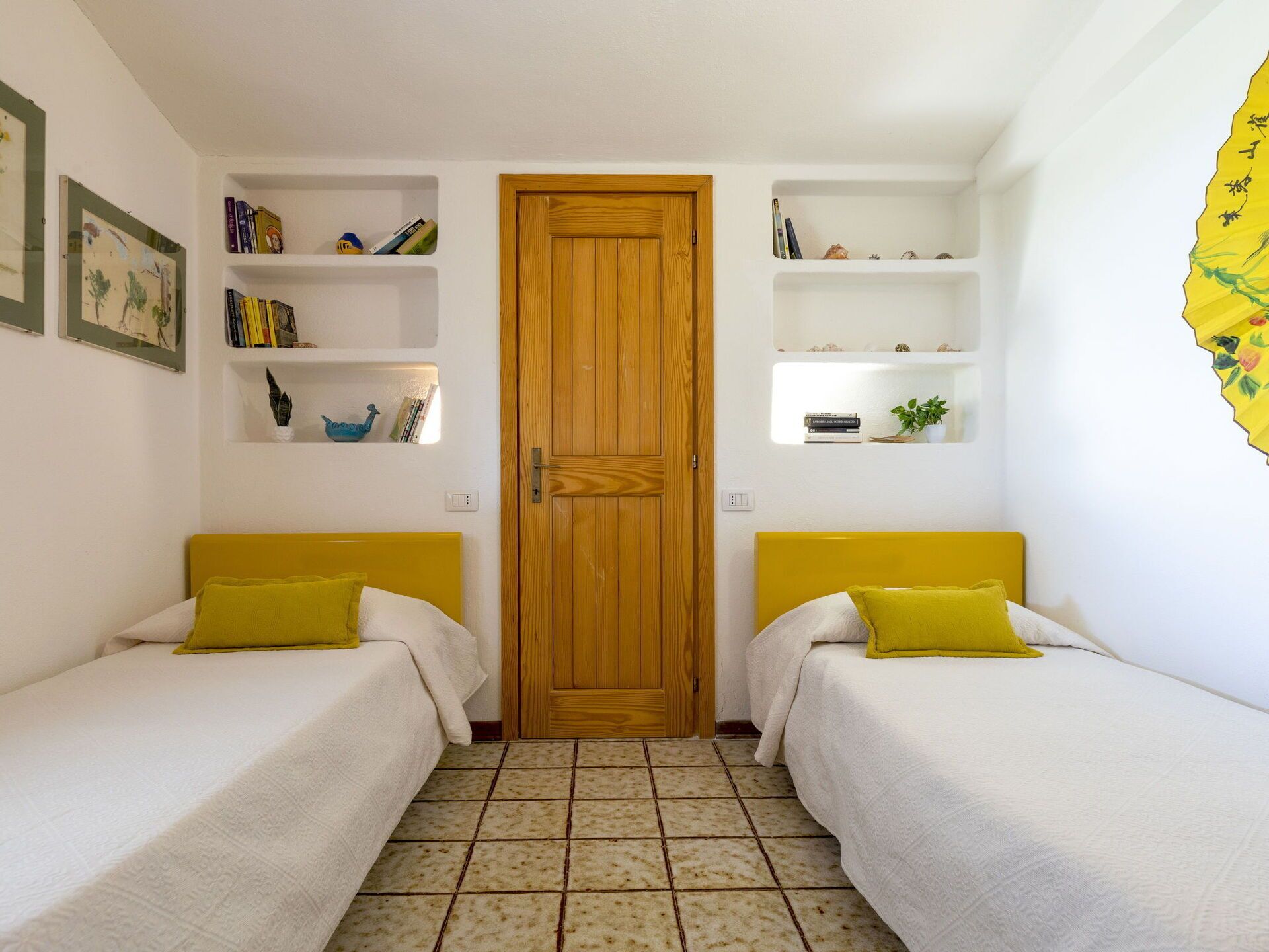 Een kamer met twee bedden, een houten deur en witte wanden met planken vol boeken en decoratieve objecten.