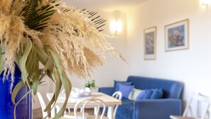 A living room with a blue sofa, a dining table with chairs, and a vase with dried plants on a counter.