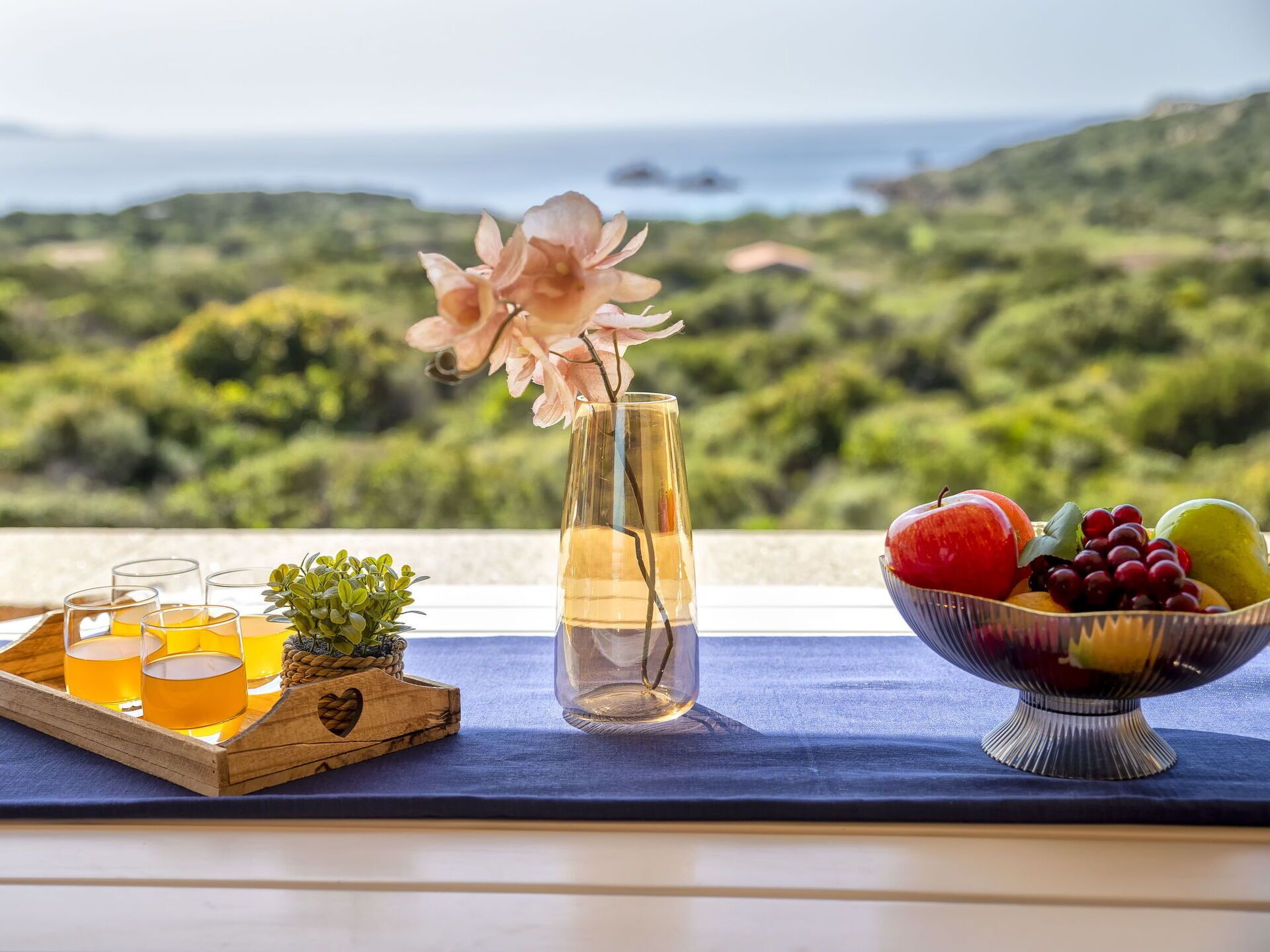 Ein Tisch mit Obstschale, Gläsern eines gelben Getränks, einer Vase mit einer Blume und Blick aufs Meer.