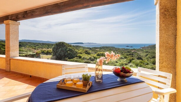 A table with a tray of drinks and fruit, a bowl of fruit, and a vase with flowers on a balcony with a view of the sea and hills.