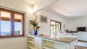 A kitchen with a window, a vase with dried plants, a white chair, and a television.