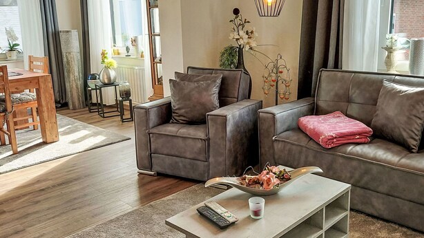A modern living room with a sofa, armchair, and coffee table. There is a dining area with a table and chairs visible through an open door. - Beautiful apartment for 2 people with WIFI, TV and terrace (Bad Pyrmont)