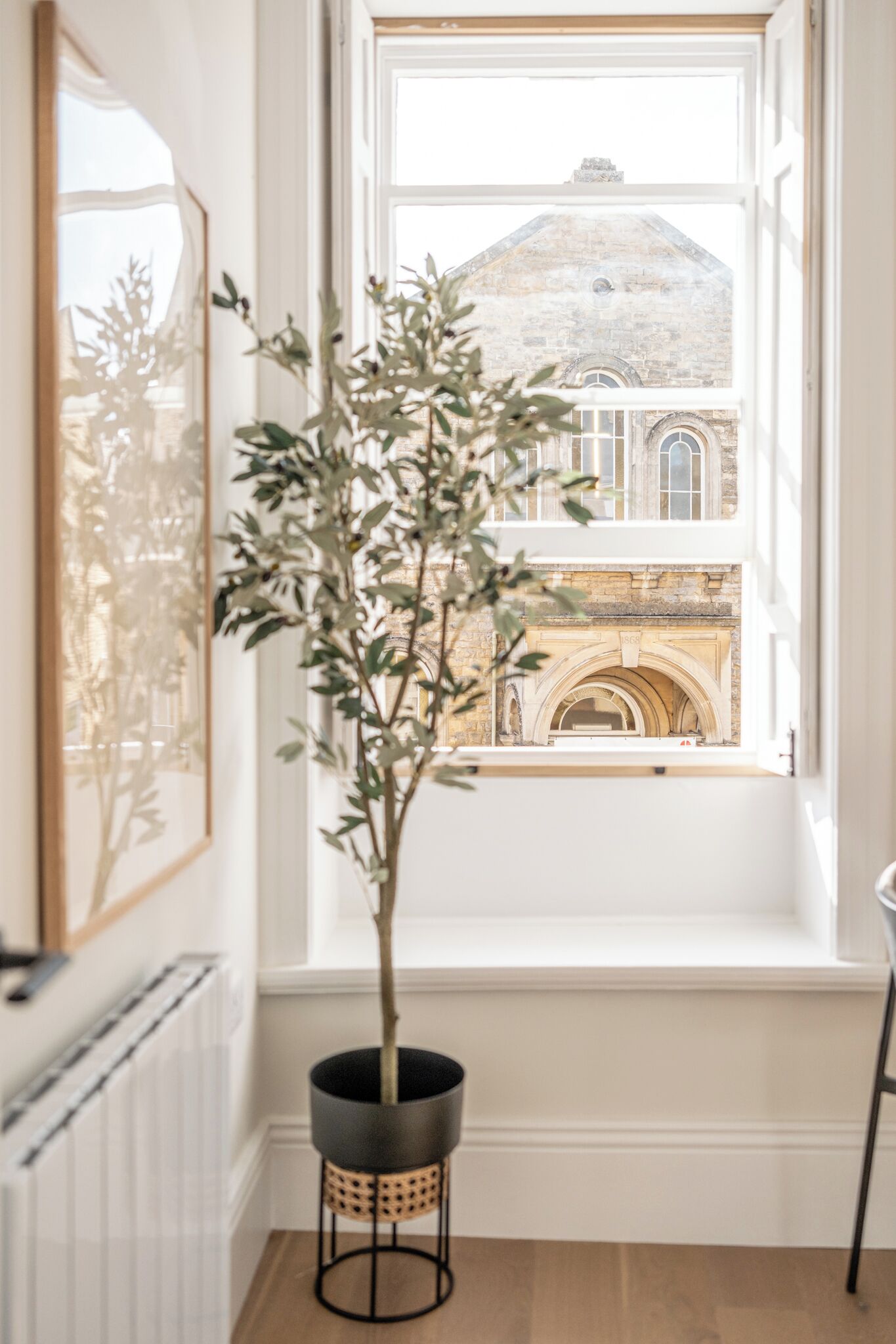 A room with a large window, a potted plant, a framed artwork, and a radiator.