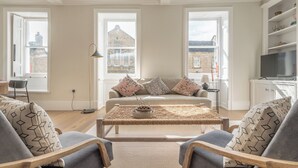 Un salón con un sofá, dos sillones, una mesa de centro y un televisor. Se ven edificios a través de dos ventanas.