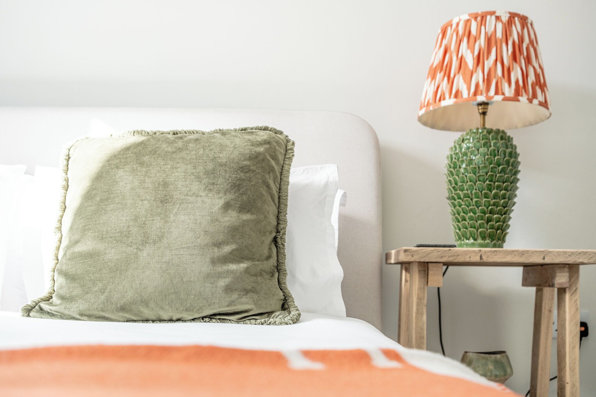 A neatly made bed with a green pillow and an orange blanket, a green cactus-shaped lamp on a wooden nightstand, and a white wall in the background.
