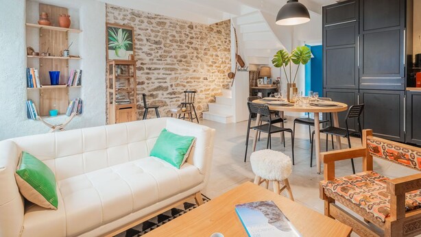 A modern living room with a white sofa, a wooden coffee table, and a dining area with a round table and chairs. There is a kitchen area with dark cabinets and a hanging light fixture. - Wonderful  villa for 5 people with WIFI and pets allowed (La Richardais)