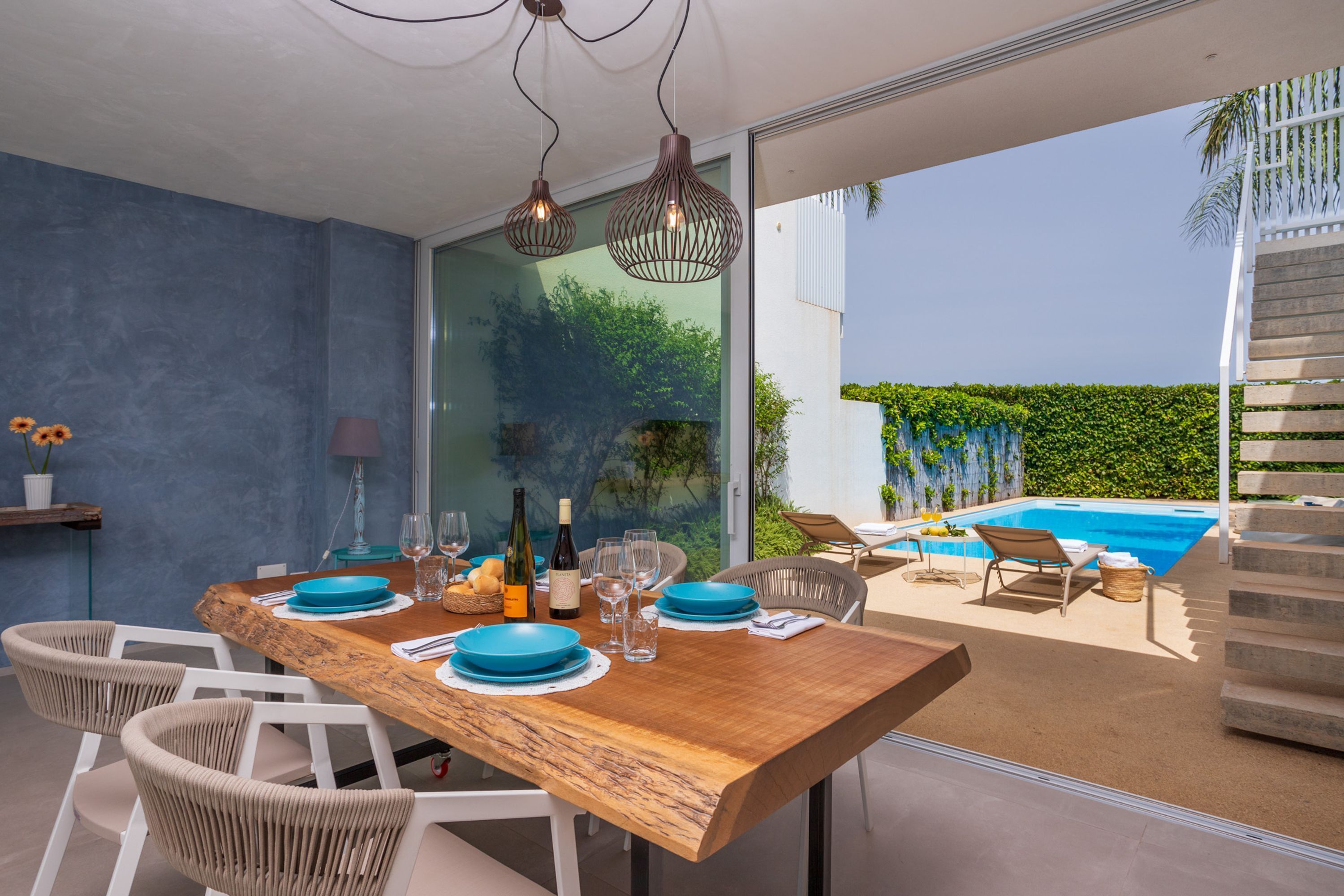Comedor con una mesa de madera dispuesta para cuatro, sillas blancas y vistas a una piscina y follaje exterior.