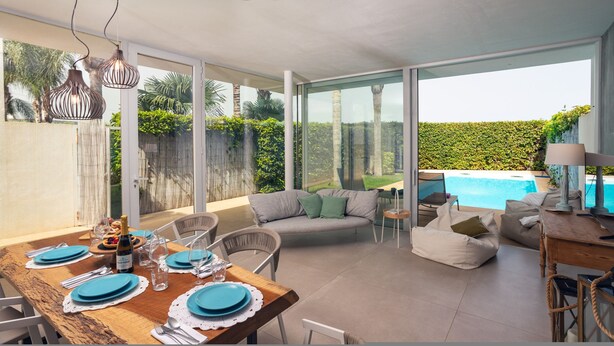 A dining area with a wooden table set for four, a sofa, and a pool view.