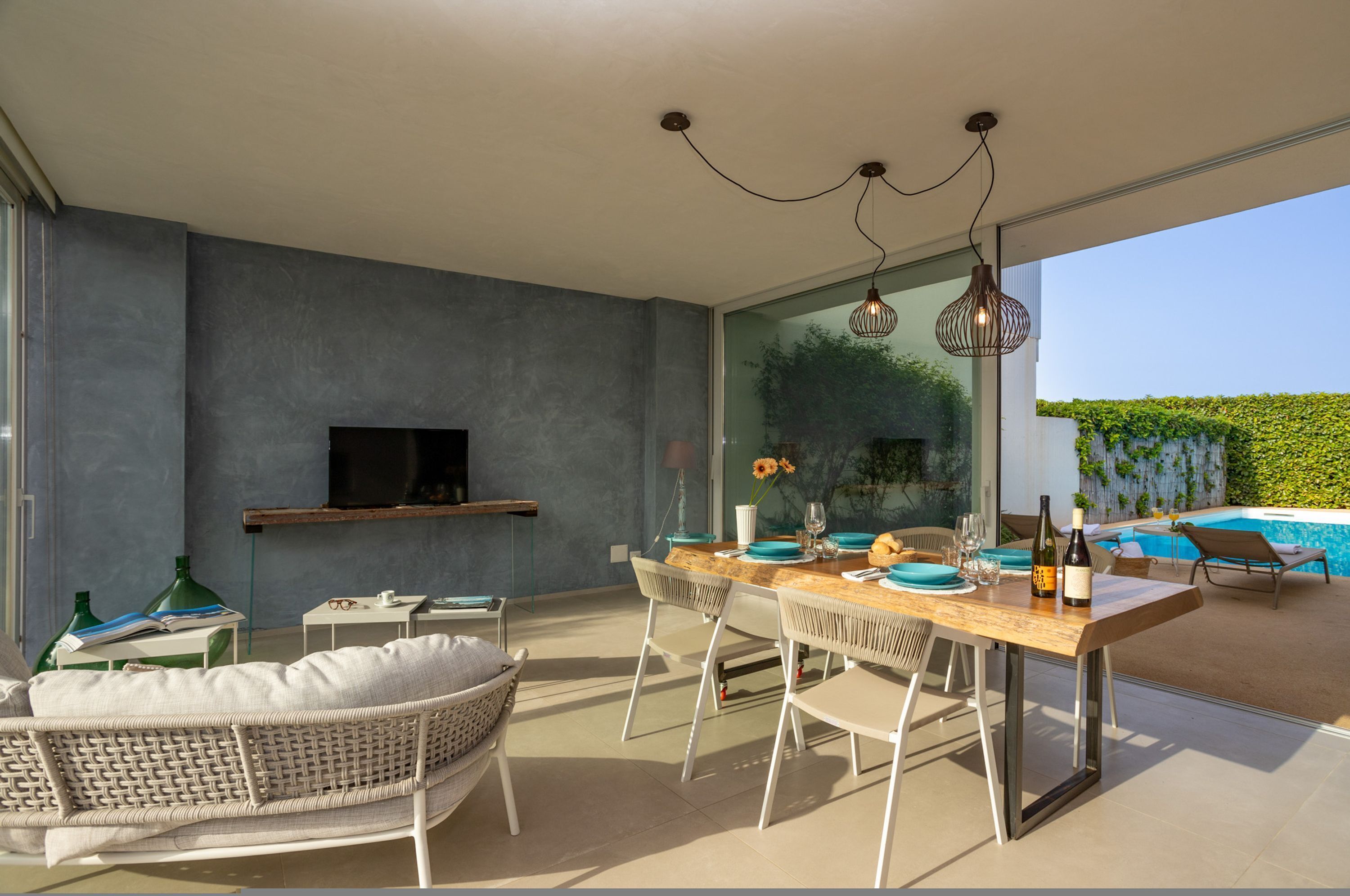 A modern outdoor dining area with a glass wall, a TV mounted on the wall, and a pool visible through the glass.
