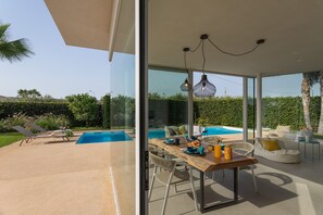 A modern outdoor dining area with a pool, lounge chairs, and a glass-enclosed patio. - Villa Mandorlo 4 Emma Villas (Ragusa)