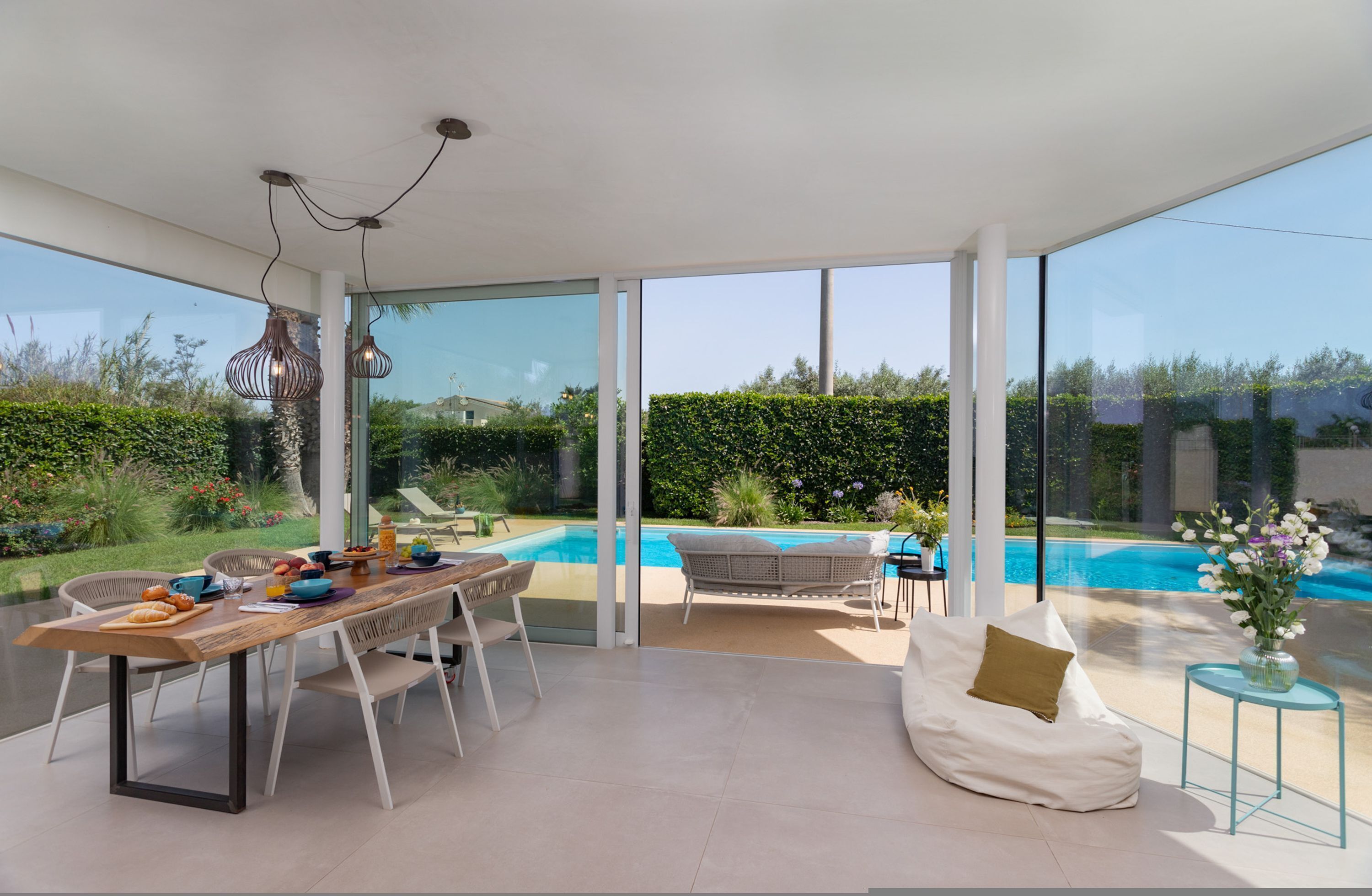 A modern outdoor dining area with a pool view, a glass-enclosed space, and a comfortable seating area.