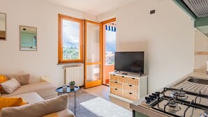 A compact living space with a sofa, a television, a kitchen area with a stove, and a view of the outdoors through a large window. - Cosy apartment for 2 people with WIFI, TV and balcony (Colico)