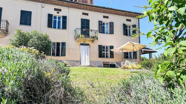 Un bâtiment à deux étages doté d’un balcon, d’une porte blanche et d’un petit jardin agrémenté de plantes et d’un parasol.