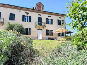 A two-story building with a balcony, a white door, and a small garden area with plants and an umbrella. - Wonderful  villa for 4 guests with WIFI and terrace (Montegrosso D'asti)