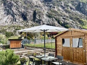 En rustik træhytte med spiseområde, et bord, stole, en parasol og udsigt til et bjerglandskab.