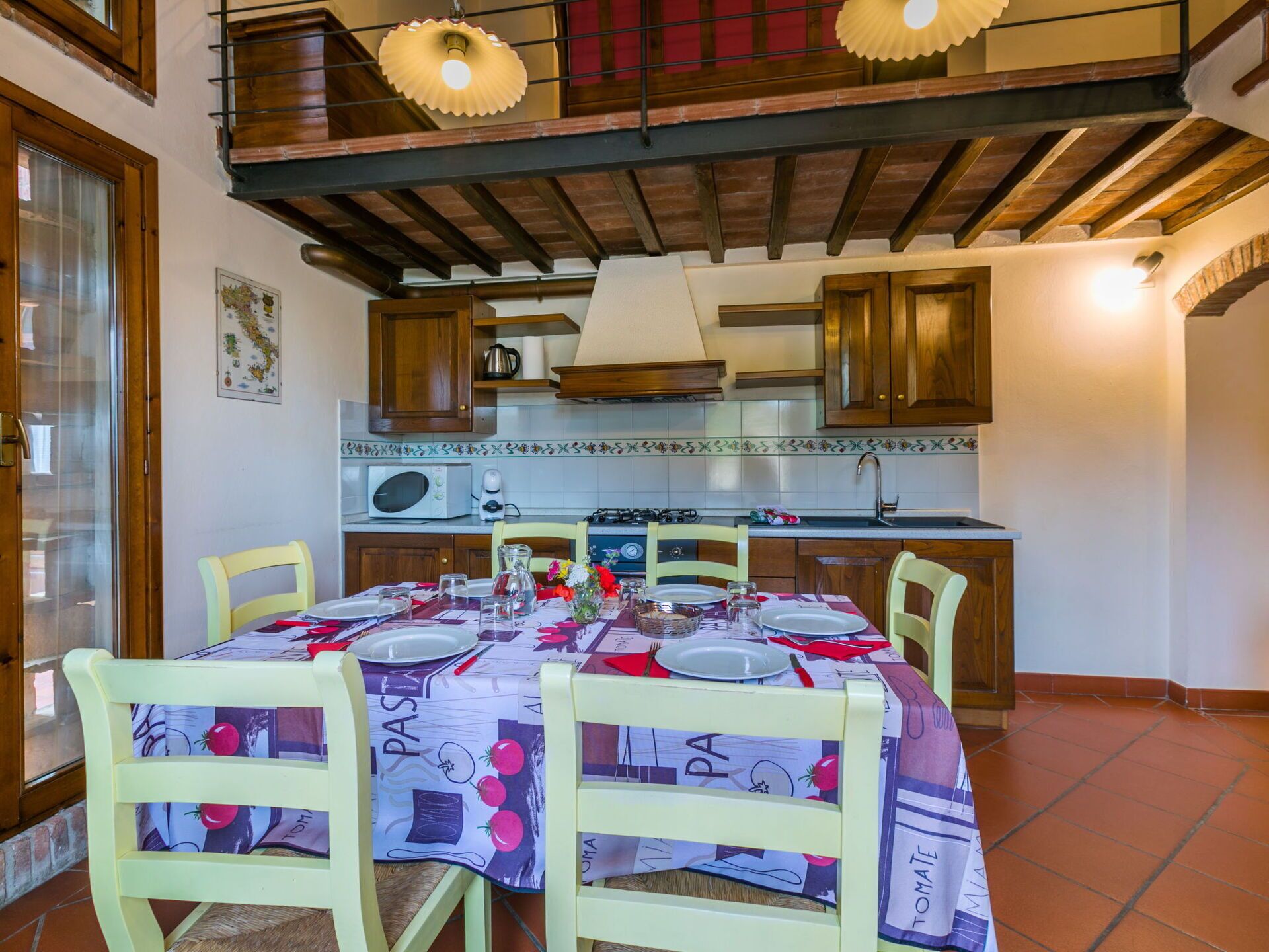 Een eetruimte met een tafel voor vier personen, omringd door houten stoelen, een keuken op de achtergrond en een balkon met houten balken.
