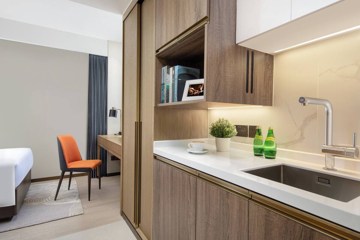 Una habitación de hotel moderna con cama, escritorio, silla, una pequeña kitchenette con pileta y una planta en maceta.