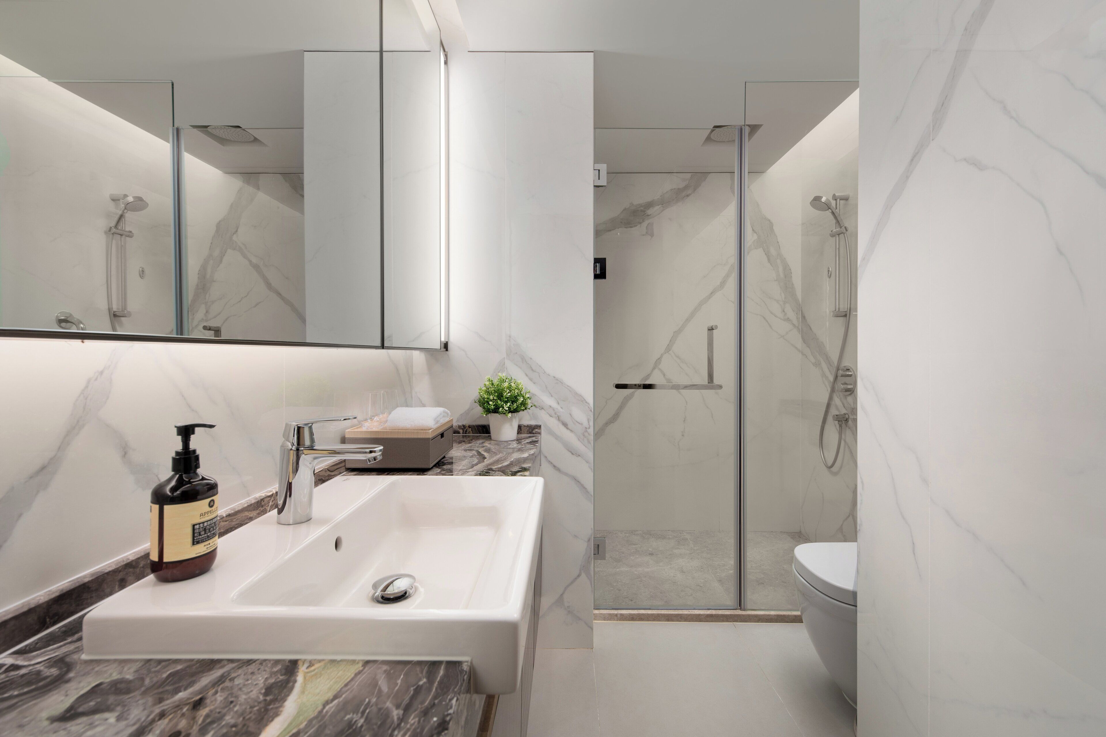 Un baño moderno con lavamanos blanco, encimera de mármol y ducha con mampara de vidrio.