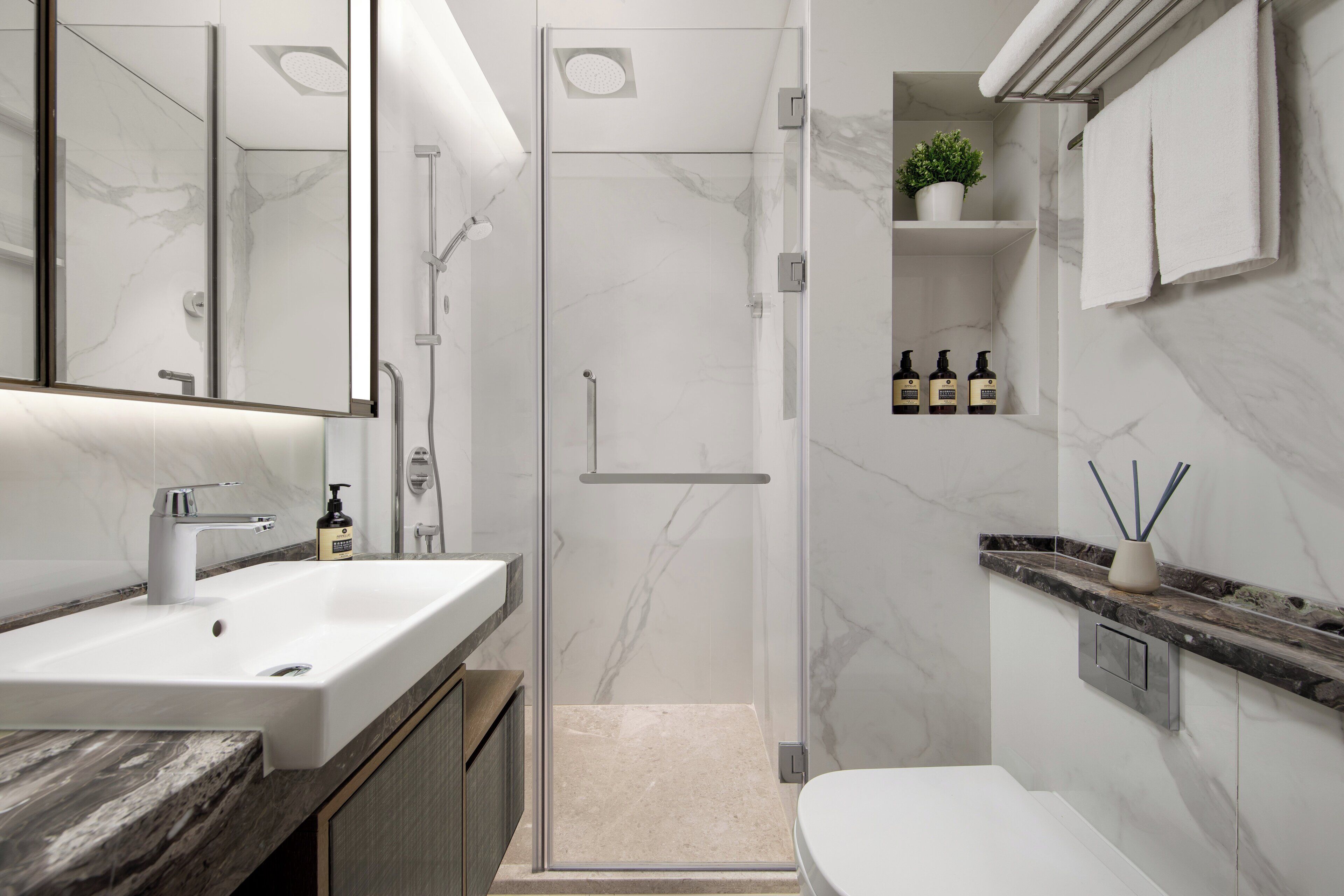 A modern bathroom with a white sink, a glass-enclosed shower, marble walls, and a small shelf with toiletries.
