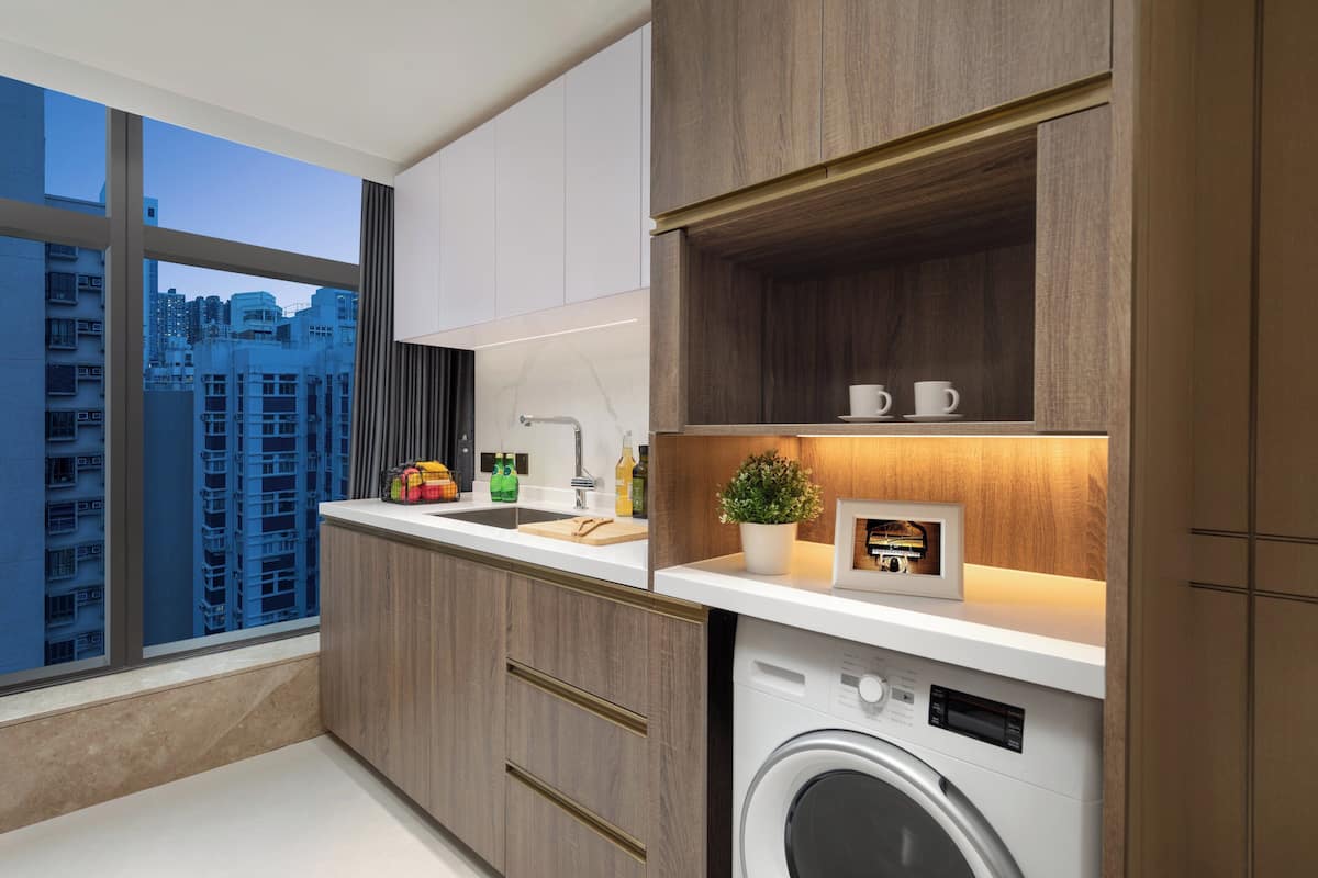 Una cocina moderna con armarios de madera, lavadora integrada y vistas a un paisaje urbano a través de amplios ventanales.