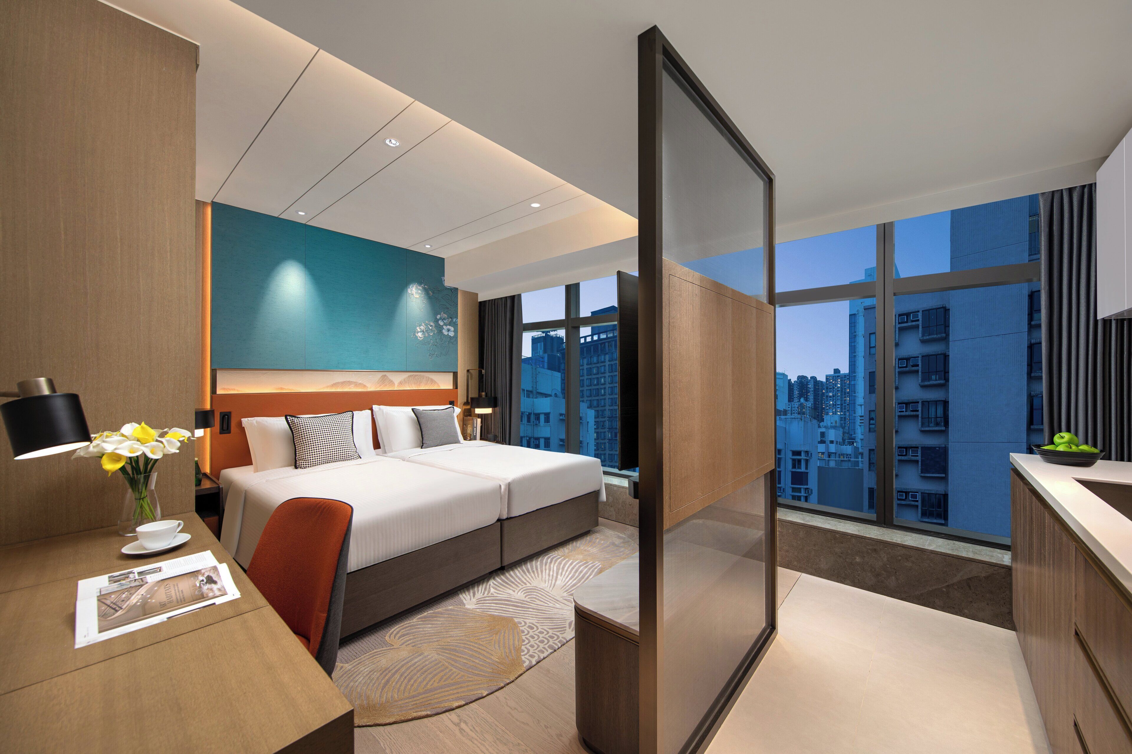 Quarto de hotel moderno com uma cama grande, uma escrivaninha com luminária, um vaso com flores e vista para a paisagem urbana através de janelas amplas.