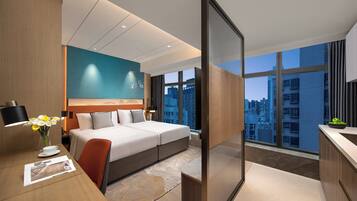 Quarto de hotel moderno com uma cama grande, uma escrivaninha com luminária, um vaso com flores e vista para a paisagem urbana através de janelas amplas.