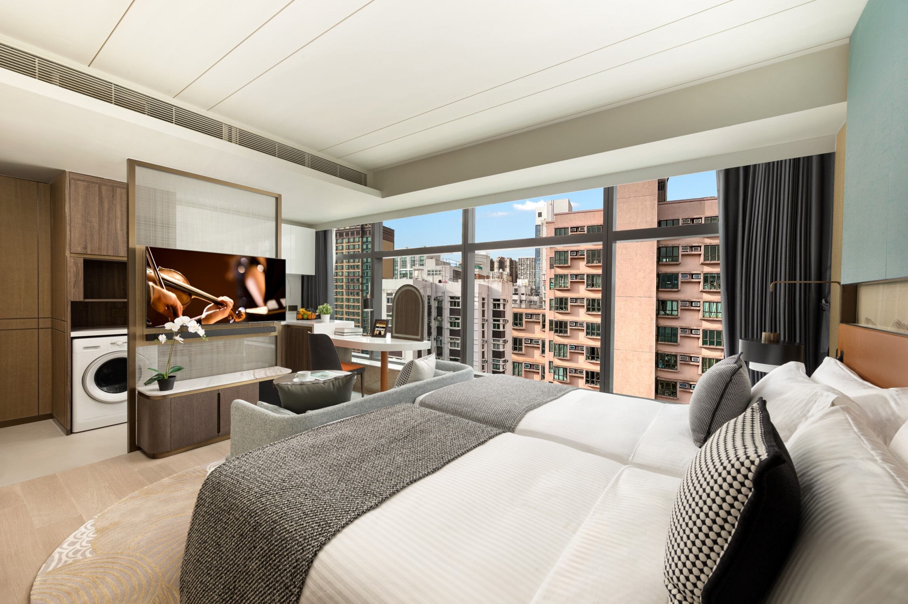 A modern hotel room with a large bed, a desk, a washing machine, and a view of high-rise buildings.
