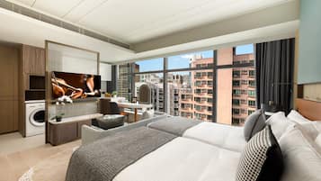 A modern hotel room with a large bed, a desk, a washing machine, and a view of high-rise buildings.