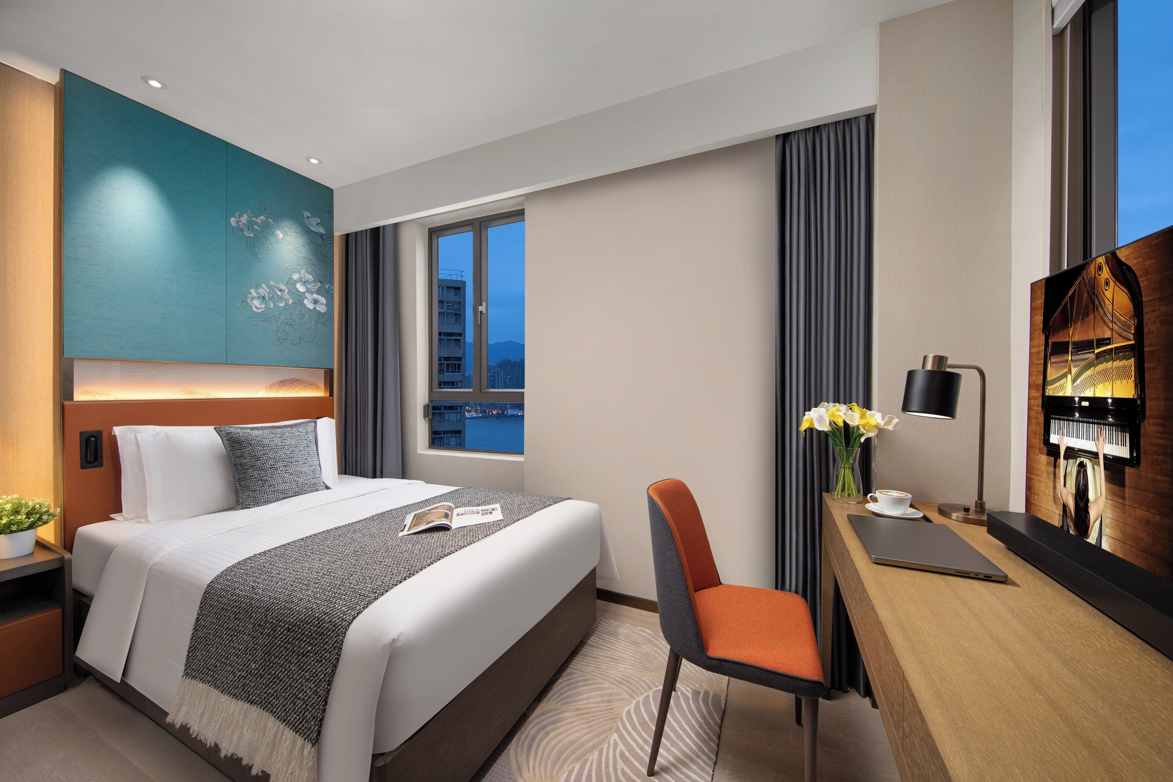 A modern hotel room with a large bed, a desk with a laptop, a chair, a vase with flowers, and a city view through the window.