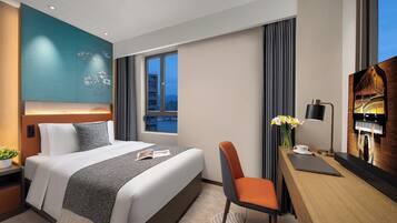 A modern hotel room with a large bed, a desk with a laptop, a chair, a vase with flowers, and a city view through the window.