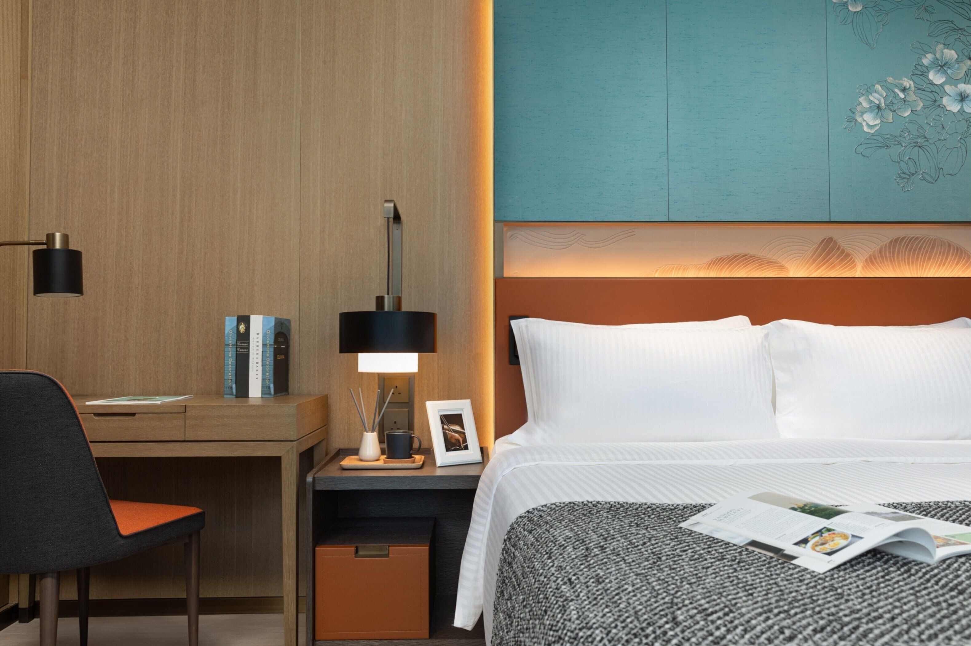 Une chambre d’hôtel avec un bureau en bois, une chaise noire, une table de chevet avec une lampe, un lit avec du linge de maison blanc et un mur orné d’un motif floral.