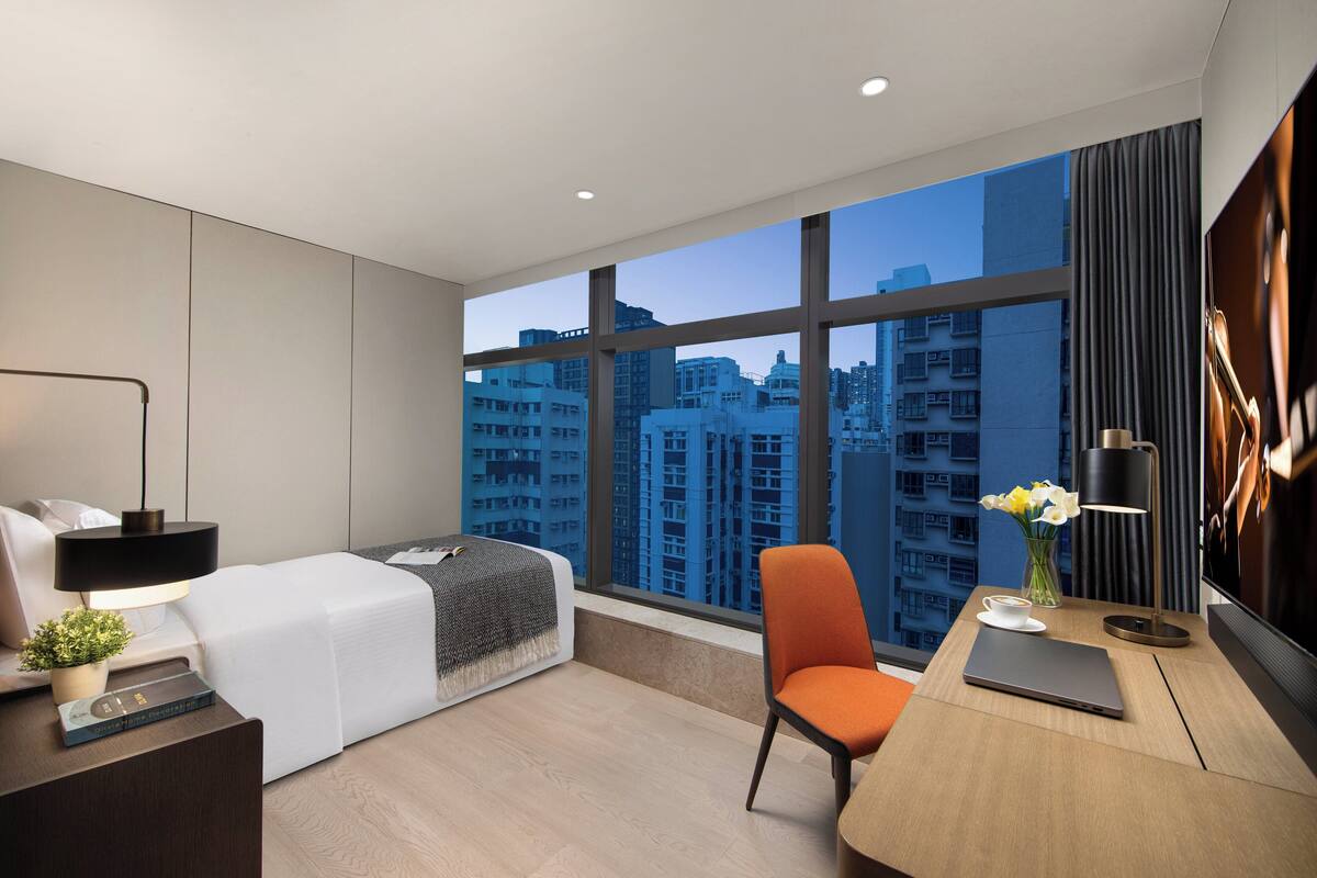 Una habitación de hotel moderna con un ventanal grande, una cama, un escritorio con una computadora portátil, una silla y una pequeña mesa con un florero.