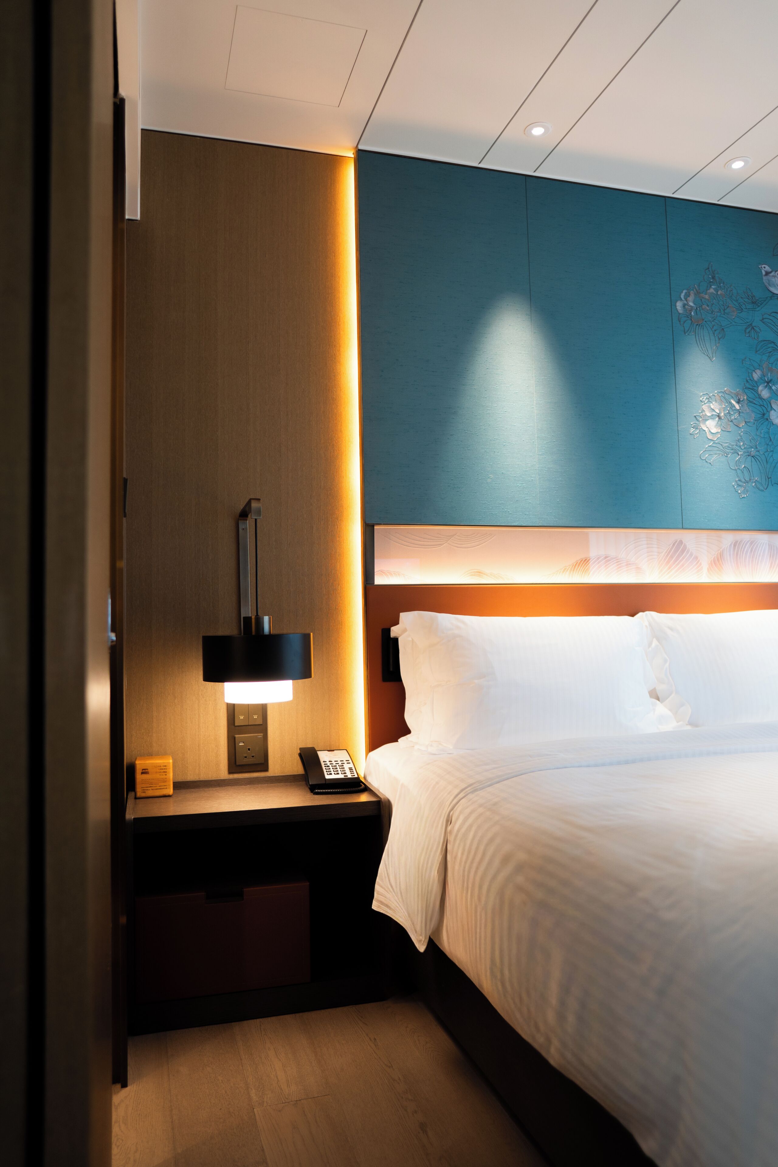 A hotel room with a bed, bedside table, lamp, and a wall with a floral design.