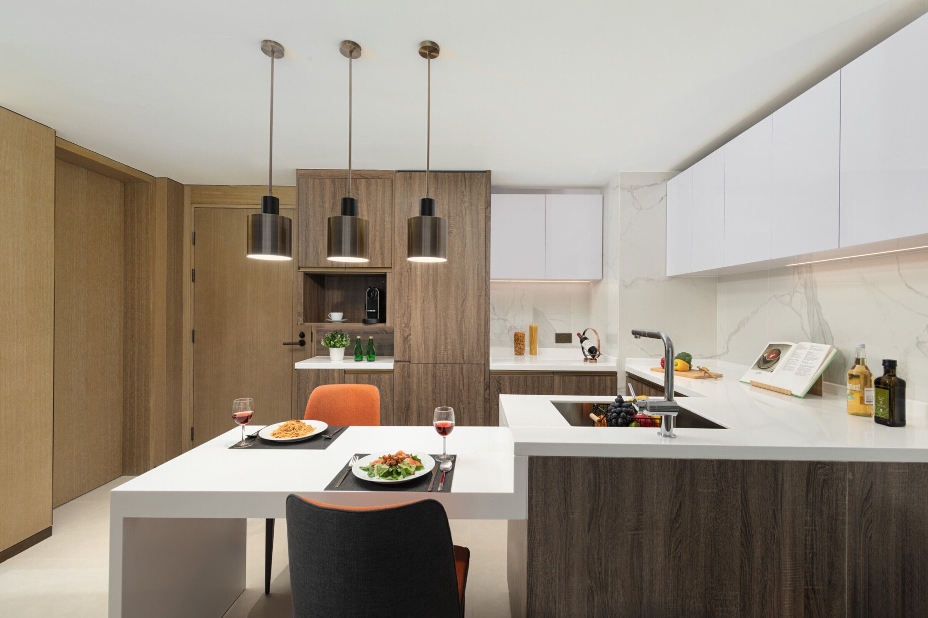 A modern kitchen with wooden cabinets, a white countertop, and pendant lighting. There is a dining table with chairs and a wine glass.