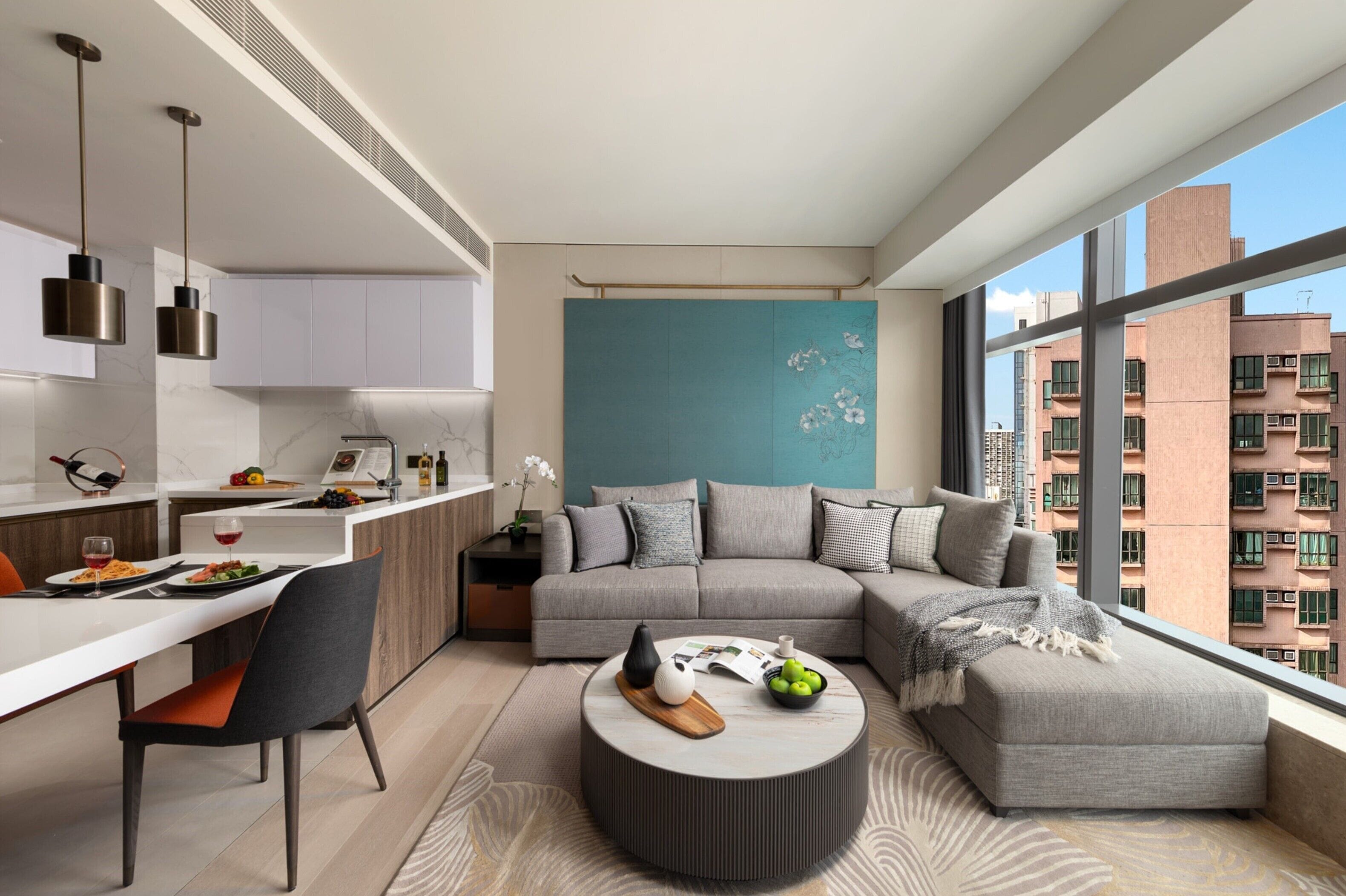 A modern apartment with a dining area, a living room with a sofa and ottoman, a kitchen with a marble countertop, and large windows offering a city view.