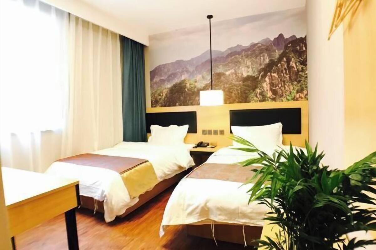 Habitación de hotel con dos camas, una mesita de noche, una lámpara, un cuadro de un paisaje montañoso y una planta en maceta.
