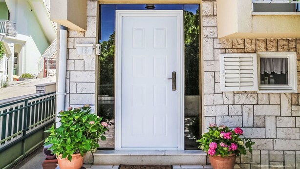 Una porta bianca con un piccolo oblò sopra, affiancata da due vasi con piante fiorite di rosa.