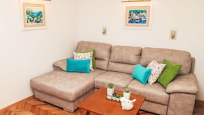 Un salón con un sofá seccional color beige, dos almohadas turquesa y verde, una mesa de centro de madera con una tetera y plantas, y dos cuadros en las paredes.