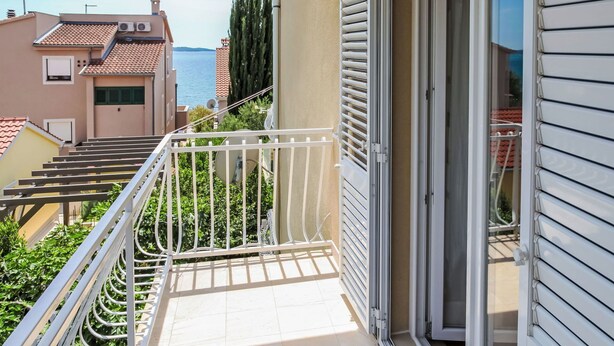 Un balcón con barandilla blanca, puerta corredera de vidrio y vista al mar y otros edificios.